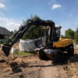 Żółto-czarna koparka Mecalac MX+ w trakcie prac ziemnych na budowie, z widocznymi elementami fundamentów i zielenią w tle.
