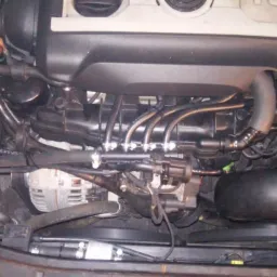 Widok z bliska na silnik samochodowy z widoczną instalacją doprowadzającą paliwo do cylindrów. Widać czarne przewody i metalowe złączki. U góry silnika widoczny napis 'TSI'.