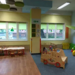 Wnętrze sali zabaw z drewnianymi meblami, stolikami, krzesłami i zabawkami w kształcie pojazdu na dywanie, widok na okna z zielonymi roletami.