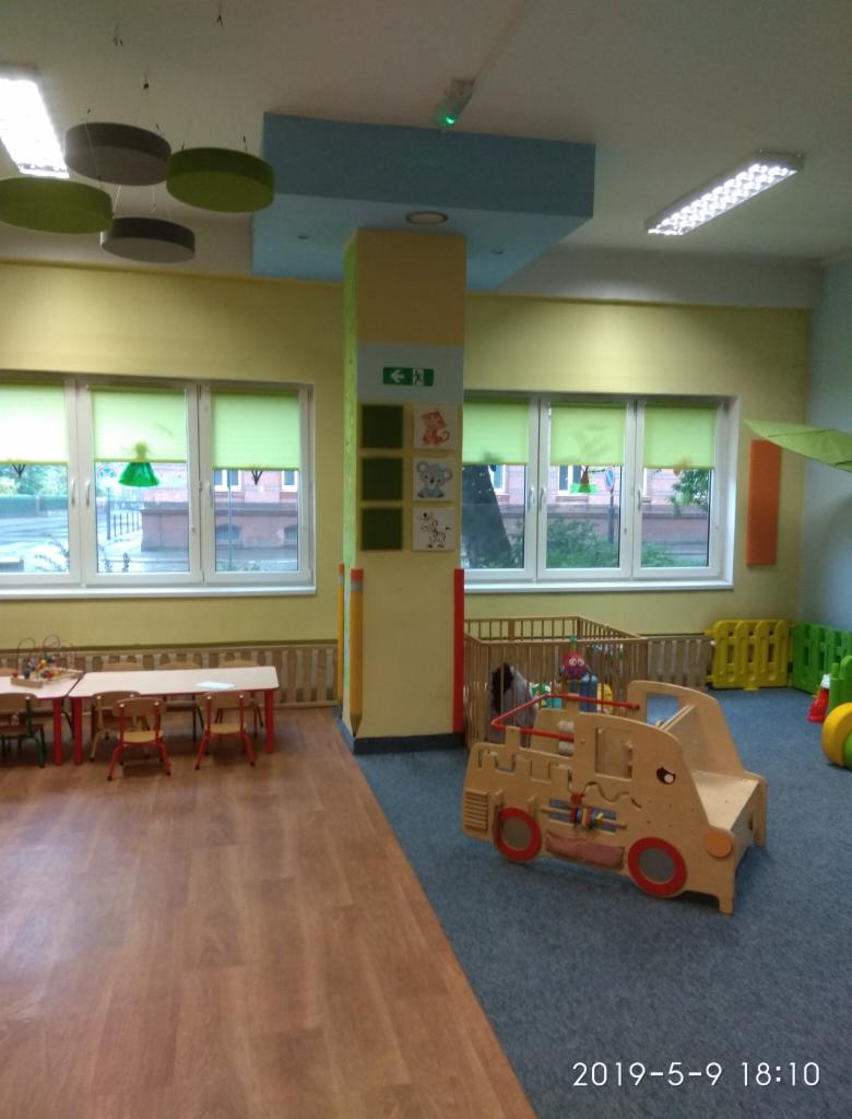 Wnętrze sali zabaw z drewnianymi meblami, stolikami, krzesłami i zabawkami w kształcie pojazdu na dywanie, widok na okna z zielonymi roletami.