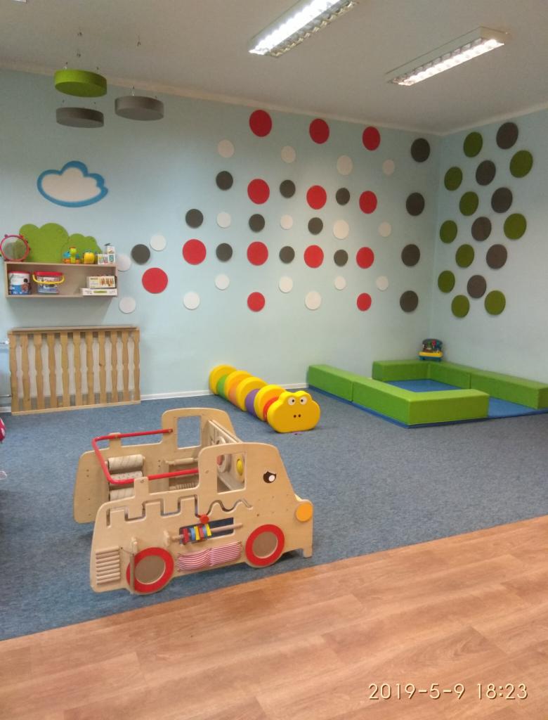 Wnętrze sali żłobkowej z drewnianym pojazdem interaktywnym, tunelem-gąsienicą, miękkimi elementami do zabawy i dekoracjami ściennymi w kropki i chmurkę.