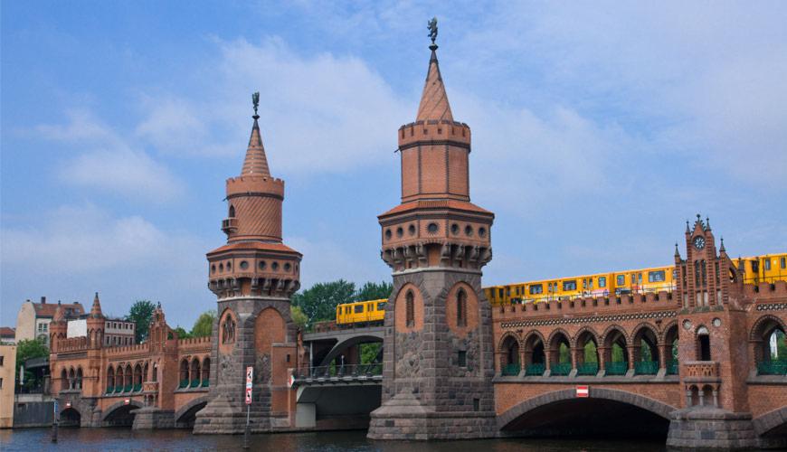 Ceglany most z dwiema wieżami zwieńczonymi posągami i żółty pociąg jadący po nim na tle błękitnego nieba.