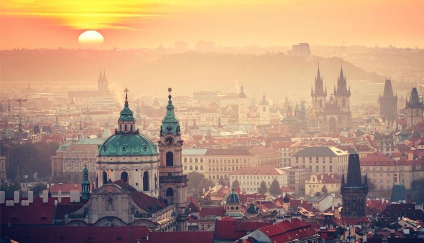 Panorama Pragi o wschodzie słońca, widok na Stare Miasto z charakterystycznymi wieżami i kopułami kościołów, delikatna mgła nad miastem.