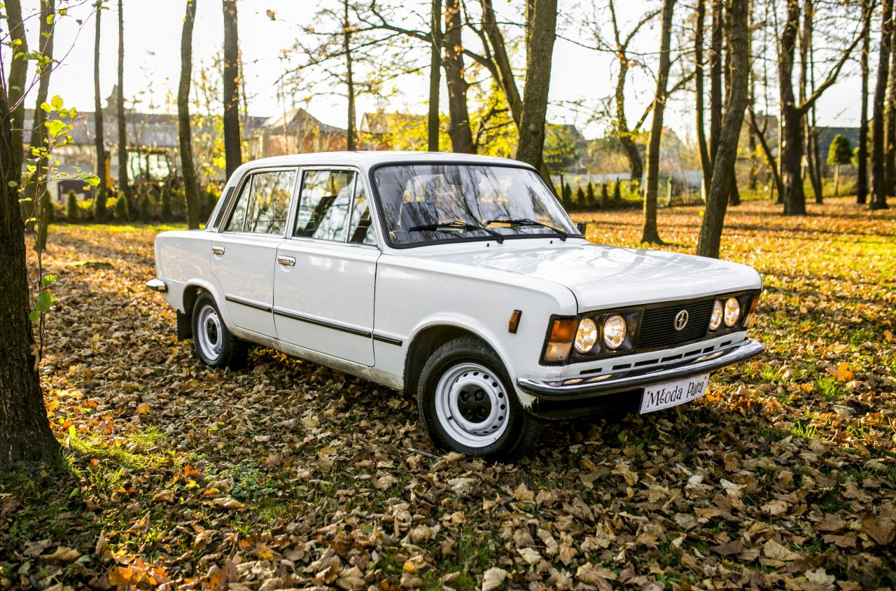 Klasyczny biały samochód osobowy FSO 125p zaparkowany na trawie pokrytej opadłymi liśćmi, w tle drzewa i budynki w jesiennym słońcu.
