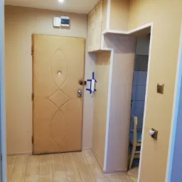 Przedpokój po remoncie: beżowe ściany, drzwi wejściowe z dekoracyjnym przeszyciem, podłoga wyłożona płytkami imitującymi drewno, widoczna szafka wisząca z jasnego drewna i fragment łazienki z białą...