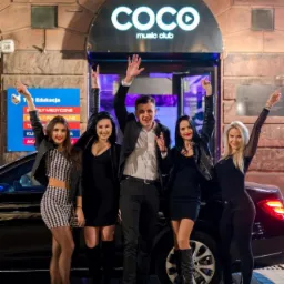 Grupa pięciu os&oacute;b, w tym jedna osoba w garniturze, pozuje z podniesionymi rękami przed wejściem do klubu nocnego 'Coco music club' z czarną limuzyną w tle.