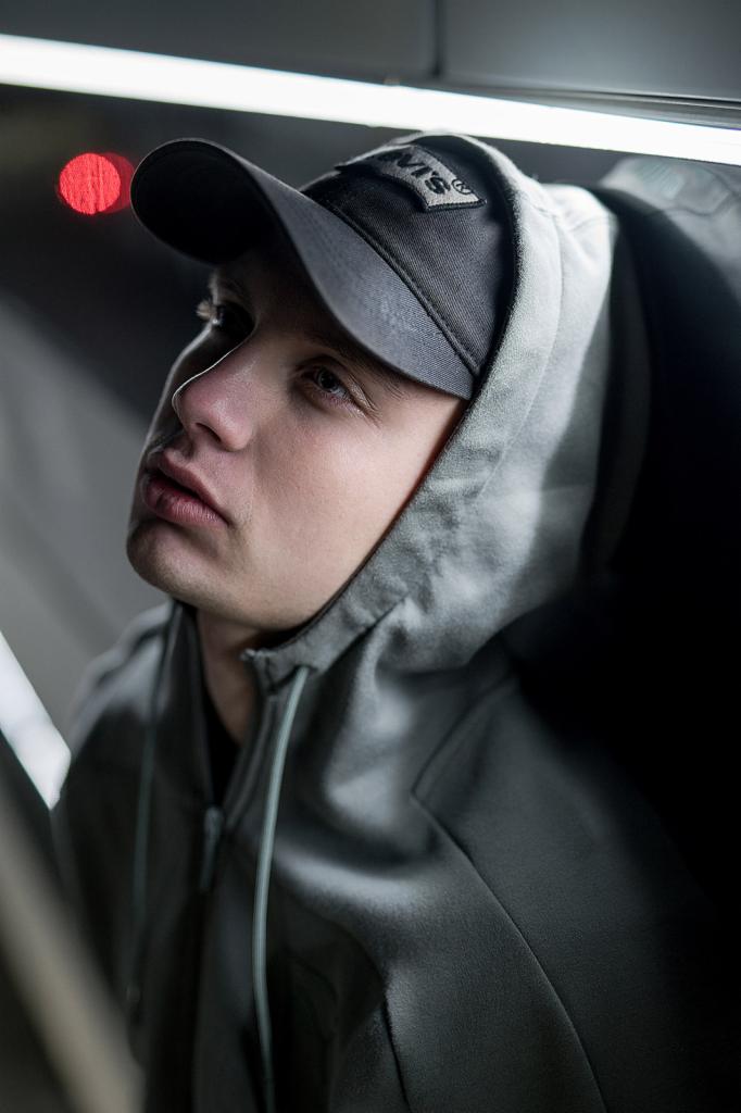 Młody mężczyzna w szarej bluzie z kapturem i czapce z daszkiem, spoglądający w górę w oświetlonym wnętrzu.