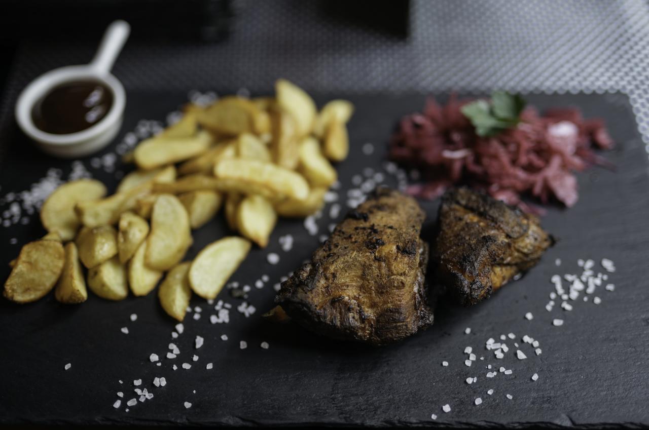 Dwa kawałki grillowanego mięsa na czarnym talerzu z frytkami w łupinkach, sosem i czerwoną kapustą posypane kryształkami soli.