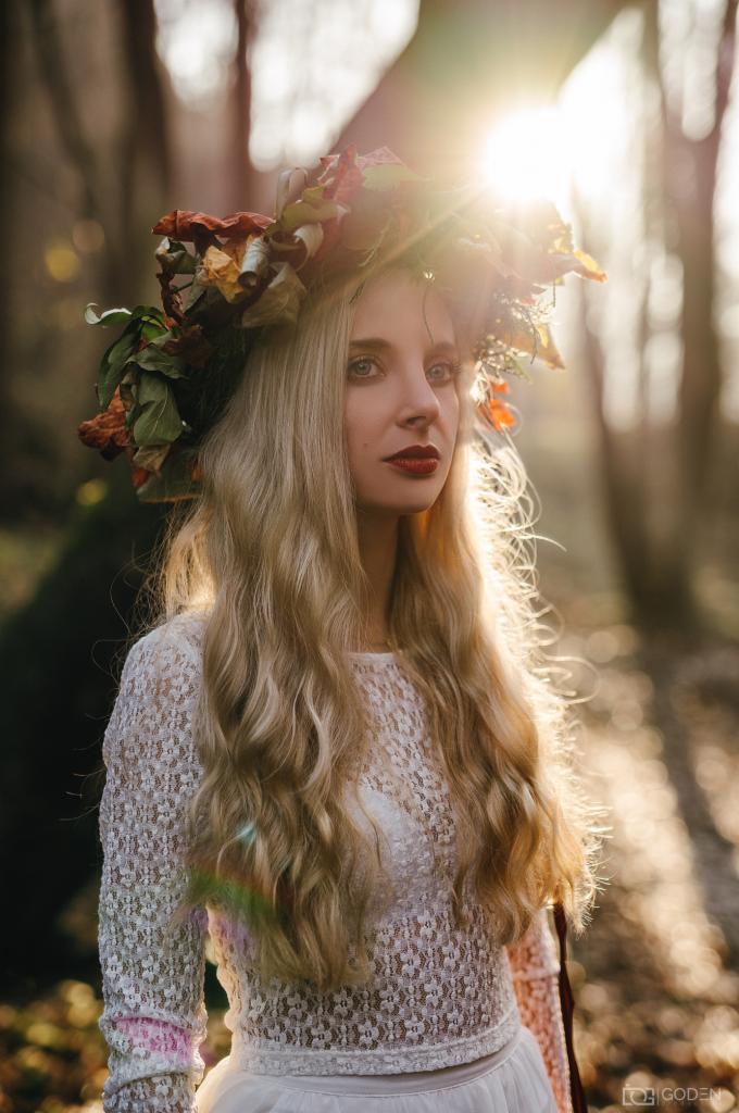 Portret kobiety w lesie z wiankiem z liści i kwiatów na głowie, w białej koronkowej bluzce, oświetlonej promieniami słońca.