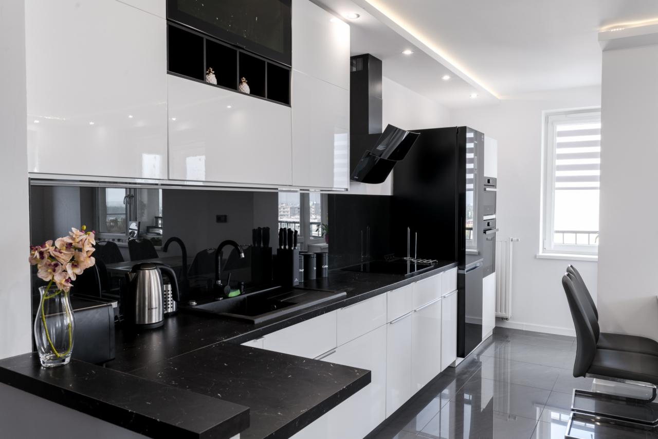 Nowoczesna kuchnia w czerni i bieli z błyszczącymi frontami szafek, czarnym blatem i szklanym panelem nad nim, odbijającym wnętrze; wazon z kwiatami na pierwszym planie.