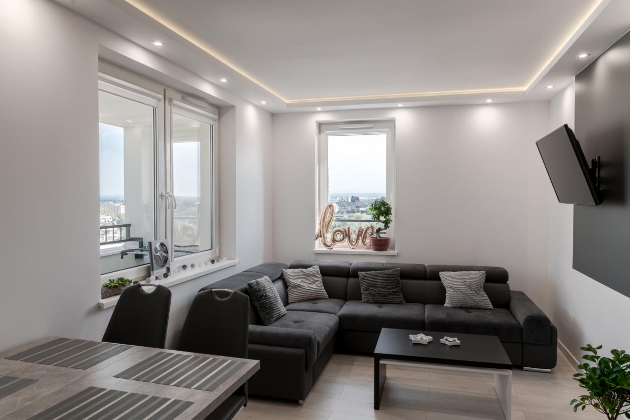 Nowoczesny salon z szarą narożną sofą, czarnym stolikiem kawowym, telewizorem na ścianie i dekoracją okna w postaci napisu 'love' wykonanego z balonów.