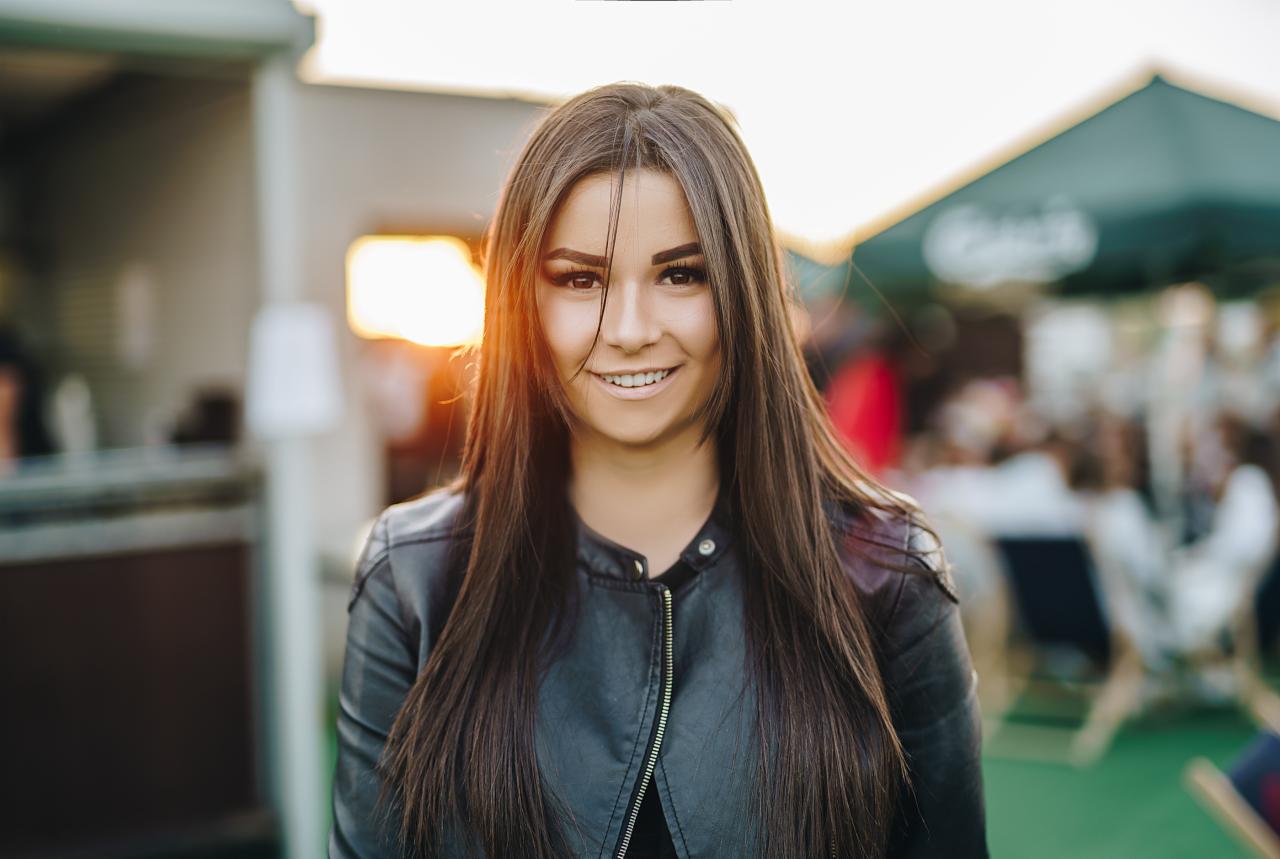 Portret uśmiechniętej kobiety w skórzanej kurtce na tle plenerowej restauracji o zachodzie słońca.