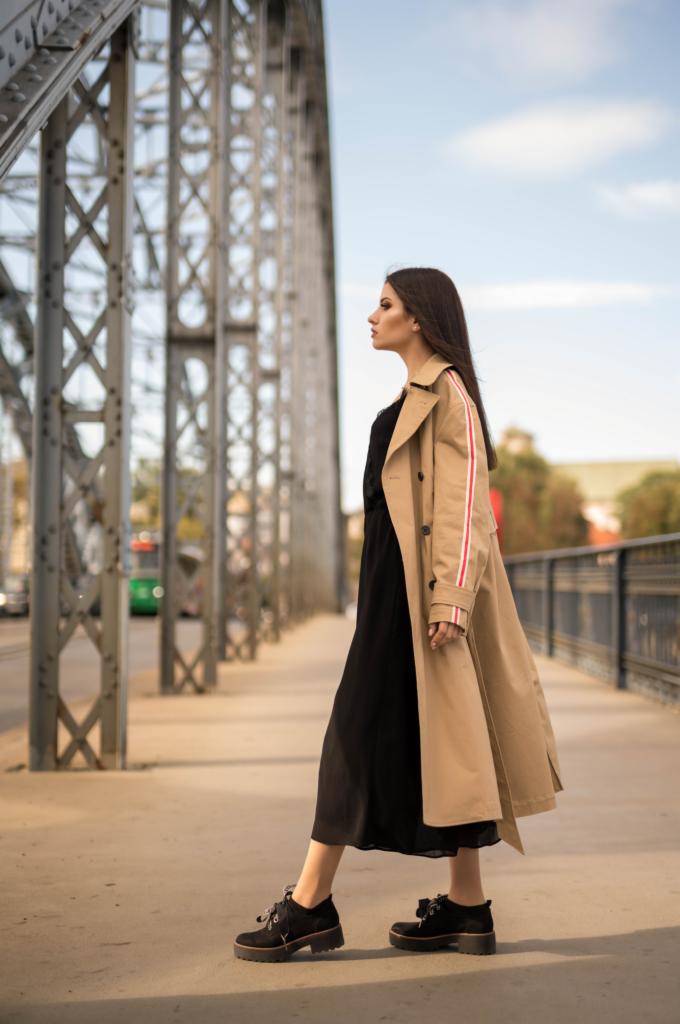 Kobieta w płaszczu z czerwonymi paskami na moście o konstrukcji kratownicowej, w tle widoczny ruch uliczny.