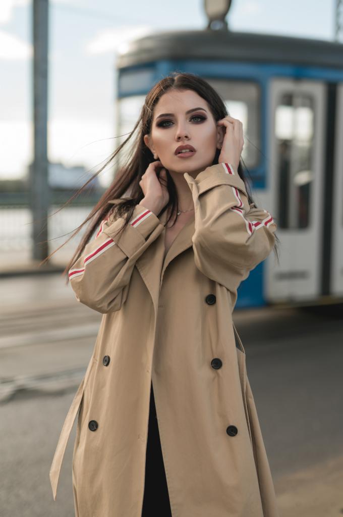 Kobieta w beżowym trenczu z czerwonymi paskami na rękawach pozuje na tle tramwaju.