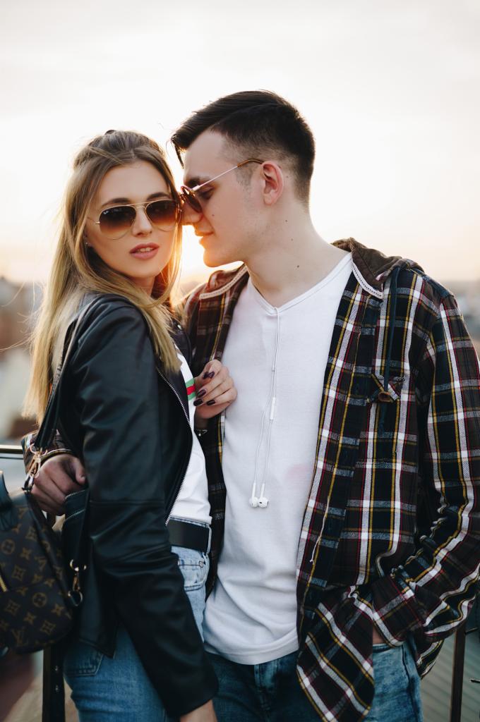 Kobieta w skórzanej kurtce i okularach przeciwsłonecznych, oparta o mężczyznę w koszuli w kratę i słuchawkach, na tle zachodzącego słońca.