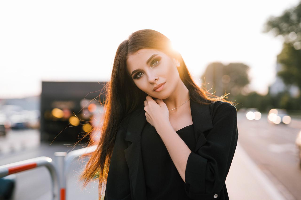 Portret kobiety w czarnym żakiecie, z delikatnym uśmiechem, oświetlonej ciepłym światłem zachodzącego słońca, z widocznymi samochodami w tle.