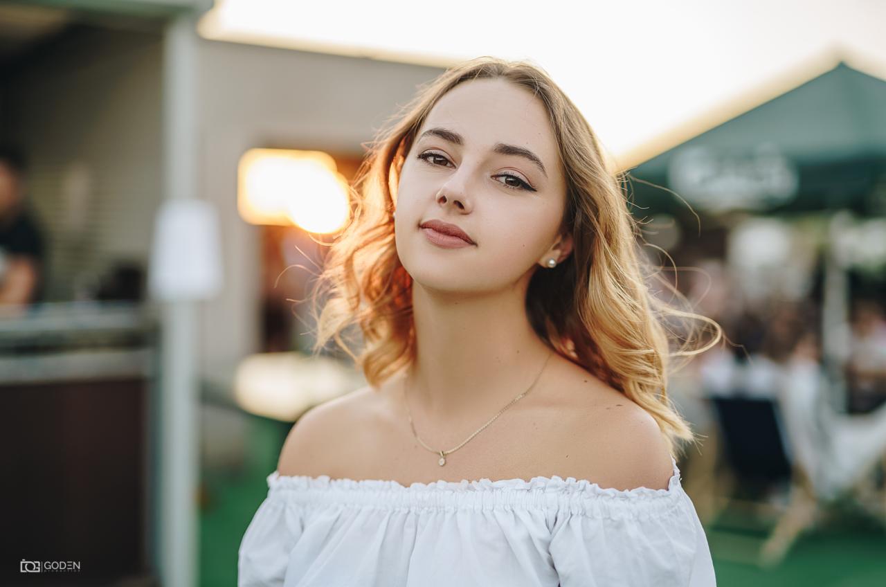 Portret młodej kobiety z blond włosami w białej bluzce hiszpance, delikatny makijaż, złoty łańcuszek, w tle rozmyte światło zachodzącego słońca i zieleń.