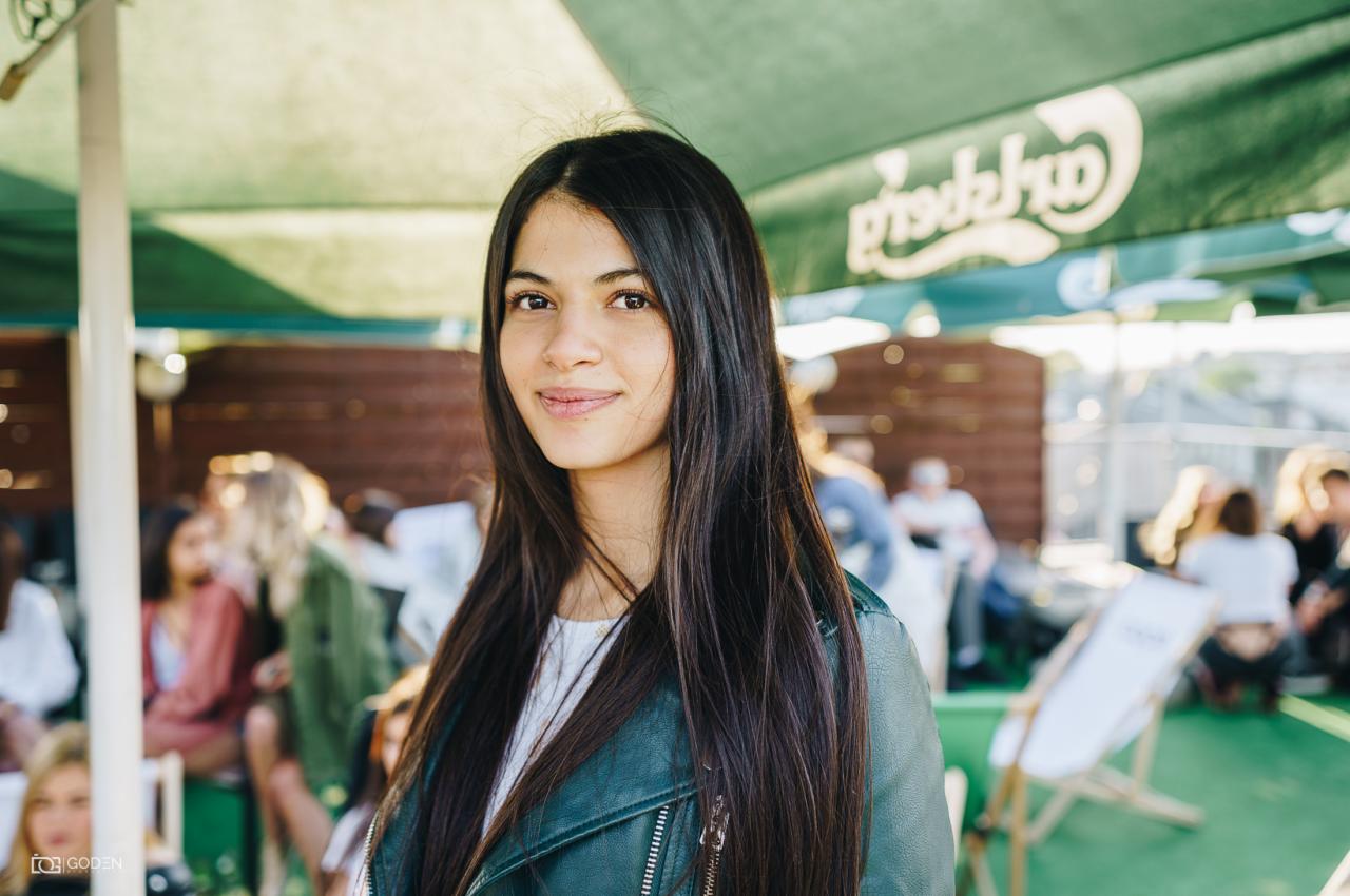 Portret uśmiechniętej brunetki w zielonej skórzanej kurtce, z widocznym logo piwa na parasolu w tle i innymi osobami w tle.