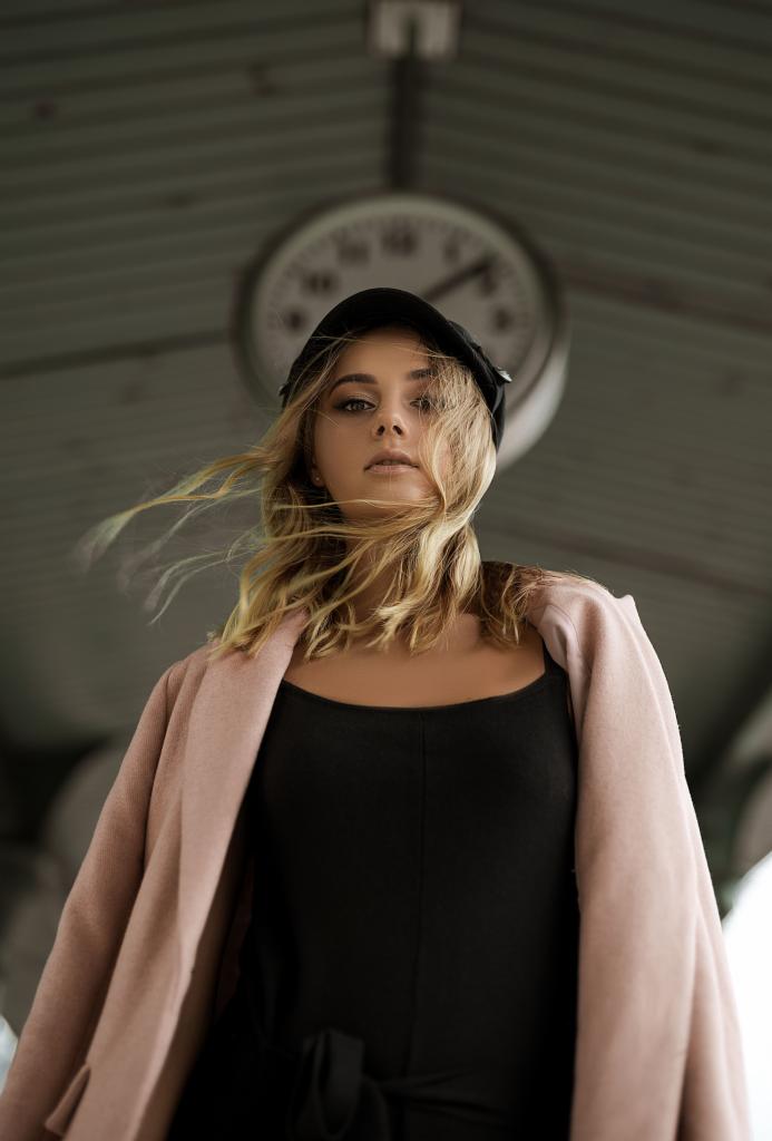 Portret kobiety w czarnej czapce i różowym płaszczu, z rozwianymi blond włosami na tle zegara.