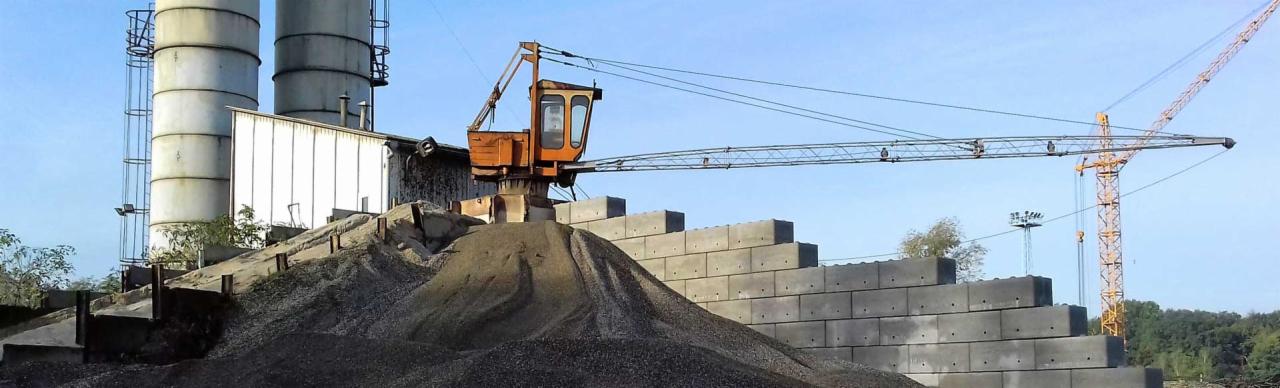 Żwirowa góra pod pomarańczowym dźwigiem na tle betonowych silosów i ściany z prefabrykatów, w tle widoczny drugi, żółty dźwig.