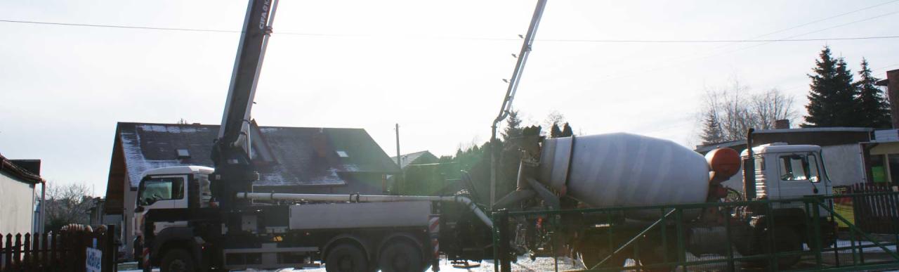 Dwa betonomieszarki na tle zabudowań mieszkalnych, jeden z wysięgnikiem pompy do betonu, w zimowej scenerii.