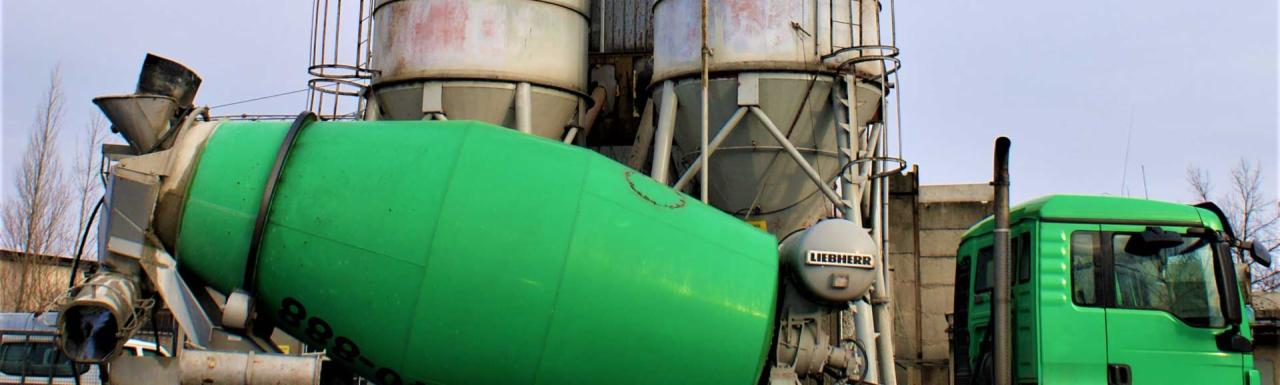 Zielona betoniarka marki Liebherr na tle silosów na składzie budowlanym, widoczny fragment kabiny ciężarówki.