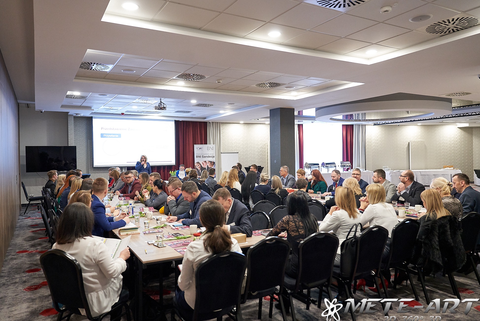 Sala konferencyjna z uczestnikami spotkania biznesowego, prelegentka prezentuje slajdy na ekranie, stoły z materiałami konferencyjnymi i napojami.
