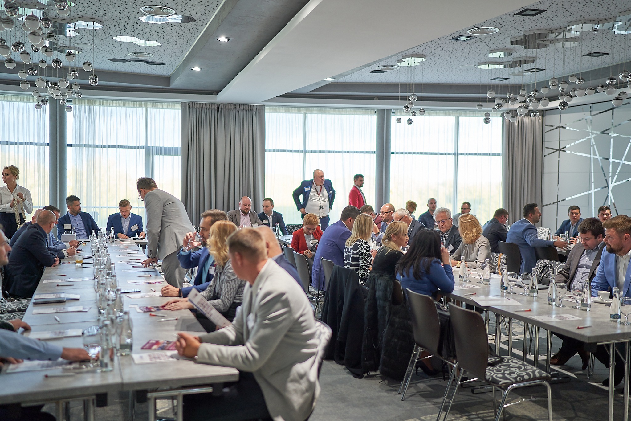 Sala konferencyjna podczas wydarzenia biznesowego, uczestnicy siedzą przy długich stołach, na stołach woda, notatki, oświetlenie sufitowe z kulistymi lampami.