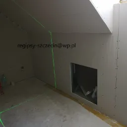 Poddasze w trakcie prac: widoczne płyty, laserowy poziom, otwór w ścianie, skos dachu z oknem.