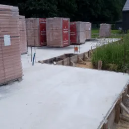 Wylana płyta fundamentowa z drewnianym szalunkiem, na tle palety z cegłami Porotherm i zieleni.