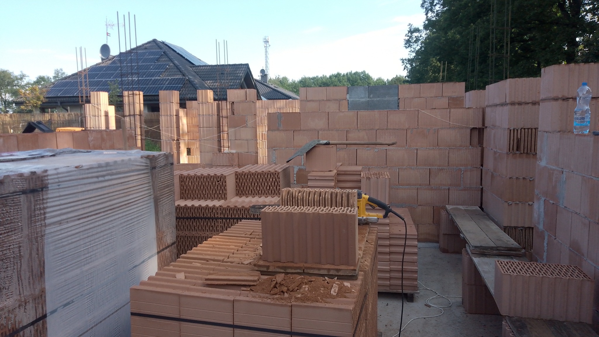 Wznoszenie ścian zewnętrznych domu z bloczków ceramicznych, widoczny murarski kielich, elektryczna bruzdownica, palety z bloczkami, dom z panelami słonecznymi w tle.