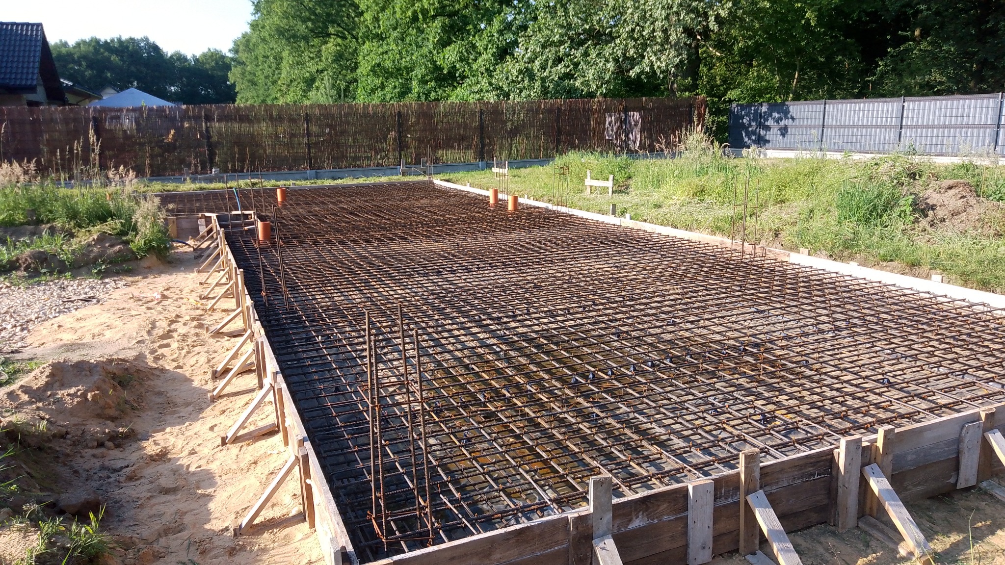 Zbrojenie płyty fundamentowej w drewnianym szalunku, widoczne pionowe pręty i pomarańczowe rury, w tle trawa i ogrodzenie.