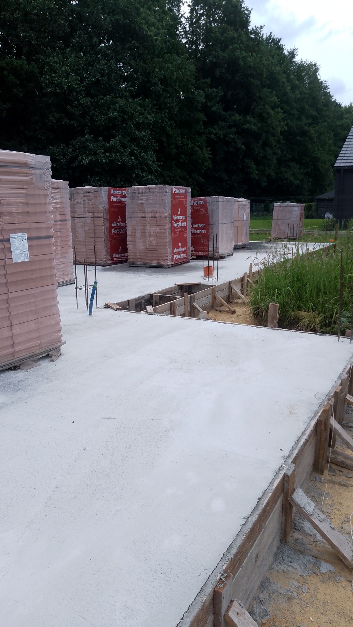 Wylana płyta fundamentowa z drewnianym szalunkiem, na tle palety z cegłami Porotherm i zieleni.