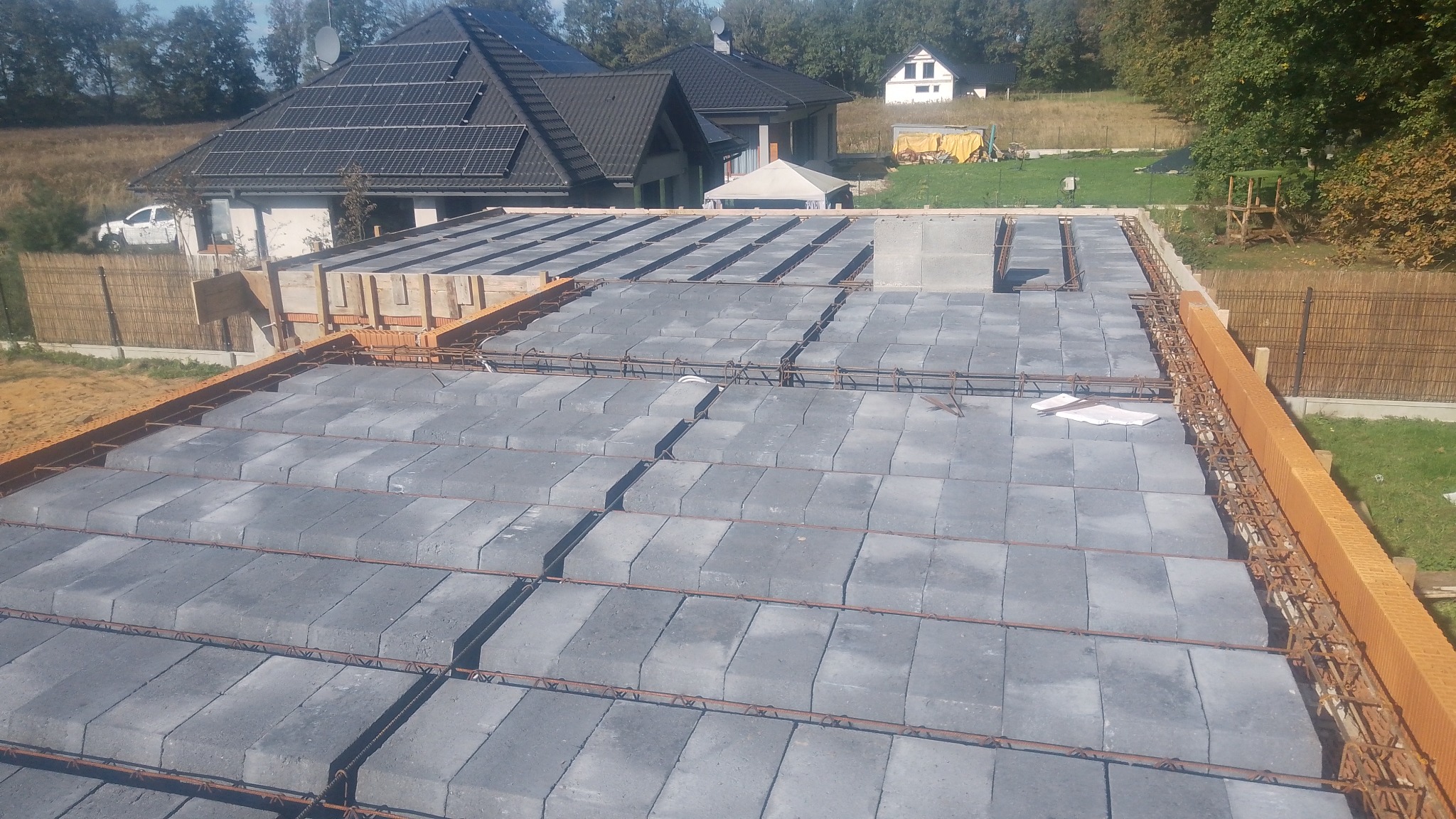 Wieniec domu w trakcie budowy: szare pustaki stropowe, zbrojenie stalowe i pomarańczowe pustaki szalunkowe, w tle dom z panelami solarnymi.