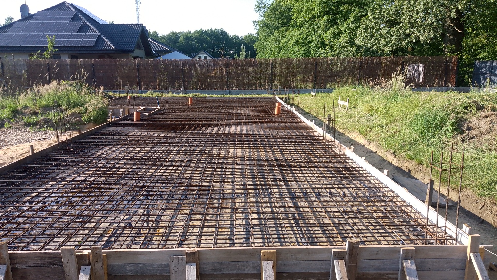 Zbrojenie fundamentu domu jednorodzinnego z drewnianym szalunkiem i wystającymi prętami pionowymi, widoczne rury kanalizacyjne, w tle dom z panelami słonecznymi i żywopłot