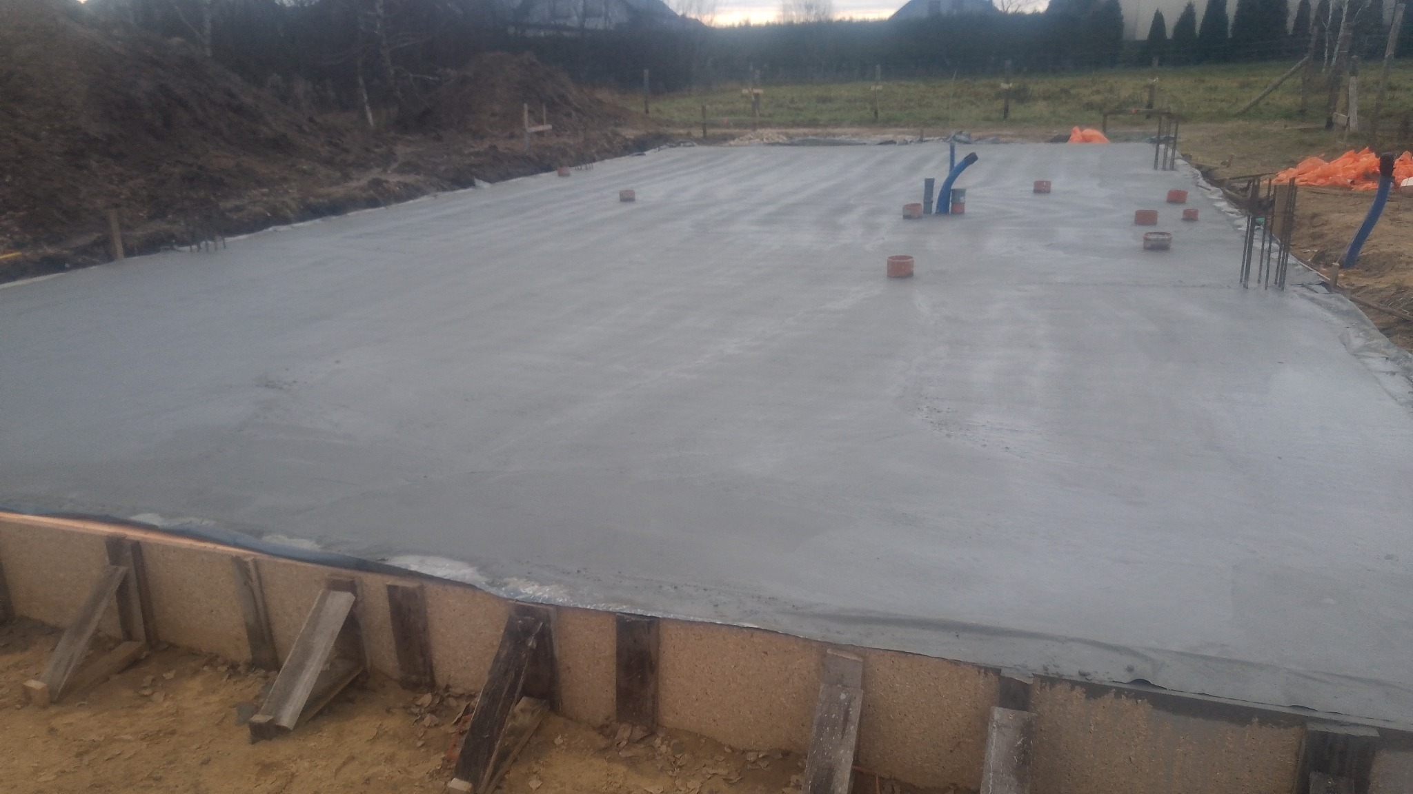 Świeżo wylana, gładka płyta fundamentowa z wystającymi rurami instalacyjnymi i prętami zbrojeniowymi, otoczona szalunkiem z desek i ziemią.
