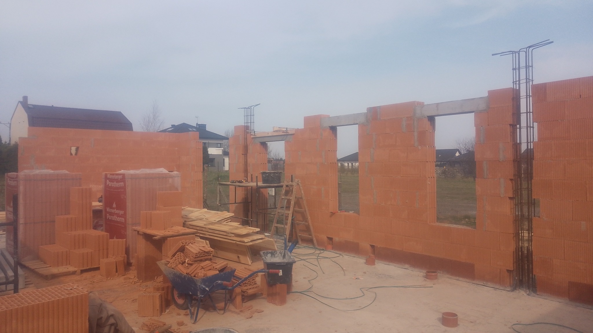 Wznoszenie ścian zewnętrznych z czerwonej cegły Porotherm, widoczne zbrojenia w narożnikach, palety z cegłami, taczka i drabina na placu budowy domu jednorodzinnego.