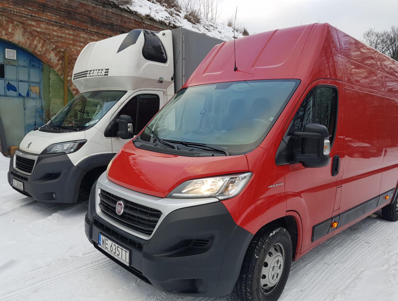 Dwa busy transportowe: biały z nadbudówką Lamar i czerwony Fiat Ducato, stojące na śniegu przed ceglanym budynkiem z niebieskimi drzwiami.