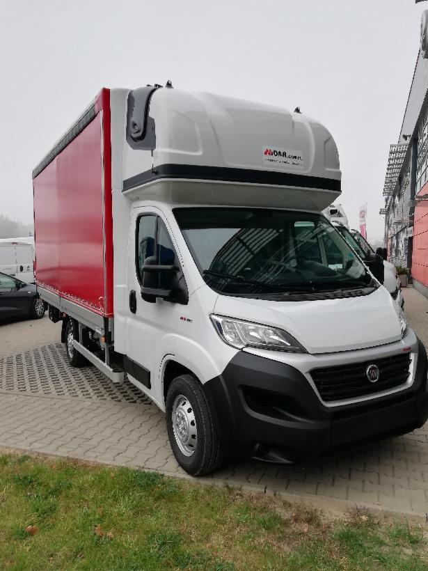 Biały bus dostawczy z czerwoną zabudową skrzyniową i podwyższonym dachem, zaparkowany na brukowanym placu przed budynkiem.