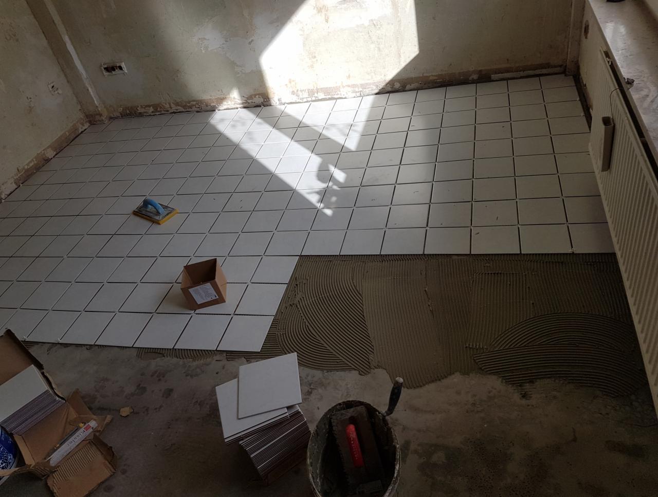 Układanie białych, kwadratowych płytek na podłodze w pomieszczeniu w trakcie remontu, widoczna warstwa kleju, narzędzia i kartony z płytkami, padające światło słoneczne.