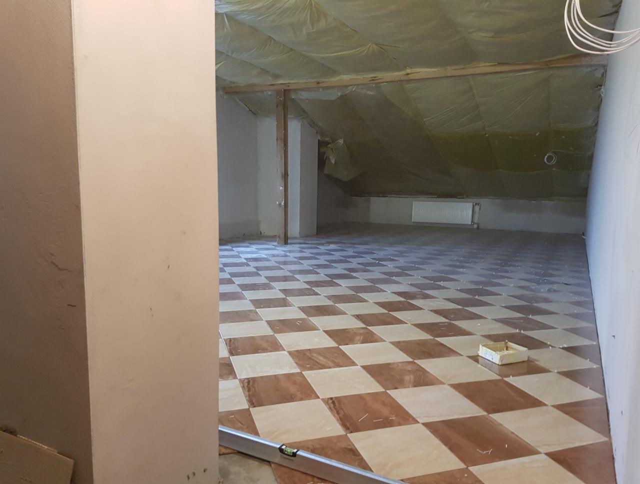 Układanie brązowo-beżowej szachownicy z płytek na poddaszu, widoczne krzyżyki dystansowe i poziomica przy ścianie.