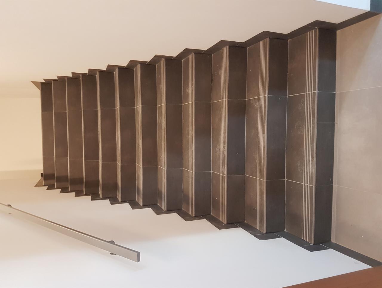 Wykończone schody z ciemnej, pionowo ułożonej glazury z jasnymi fugami w Grądy-Woniecko, widok z góry na całą długość biegu, z zamontowaną metalową poręczą.