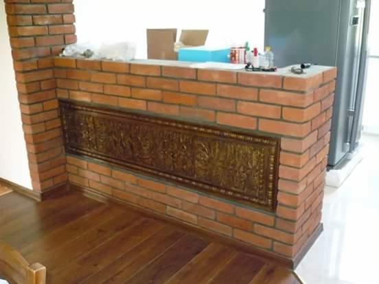 Bar z cegły klinkierowej z dekoracyjnym, ciemnym panelem z ornamentami, stojący na drewnianej podłodze, z widoczną lodówką w tle.