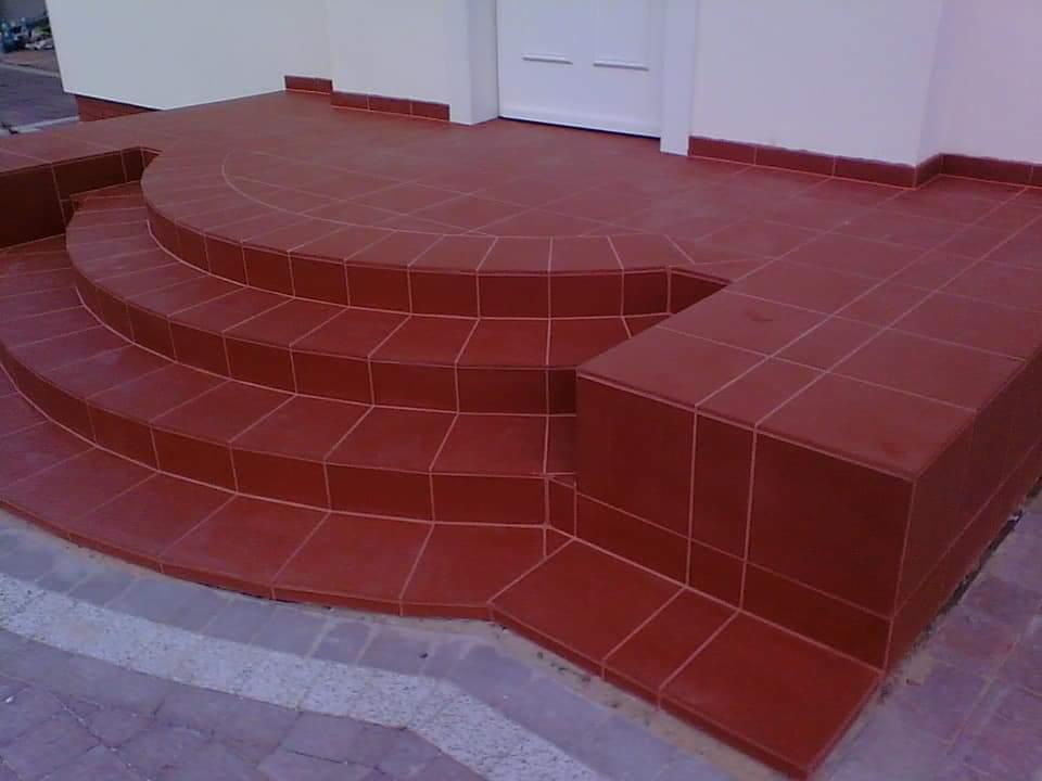 Wykończone płytkami w odcieniach czerwieni schody wejściowe do budynku, z widocznym fragmentem białej elewacji i drzwi.