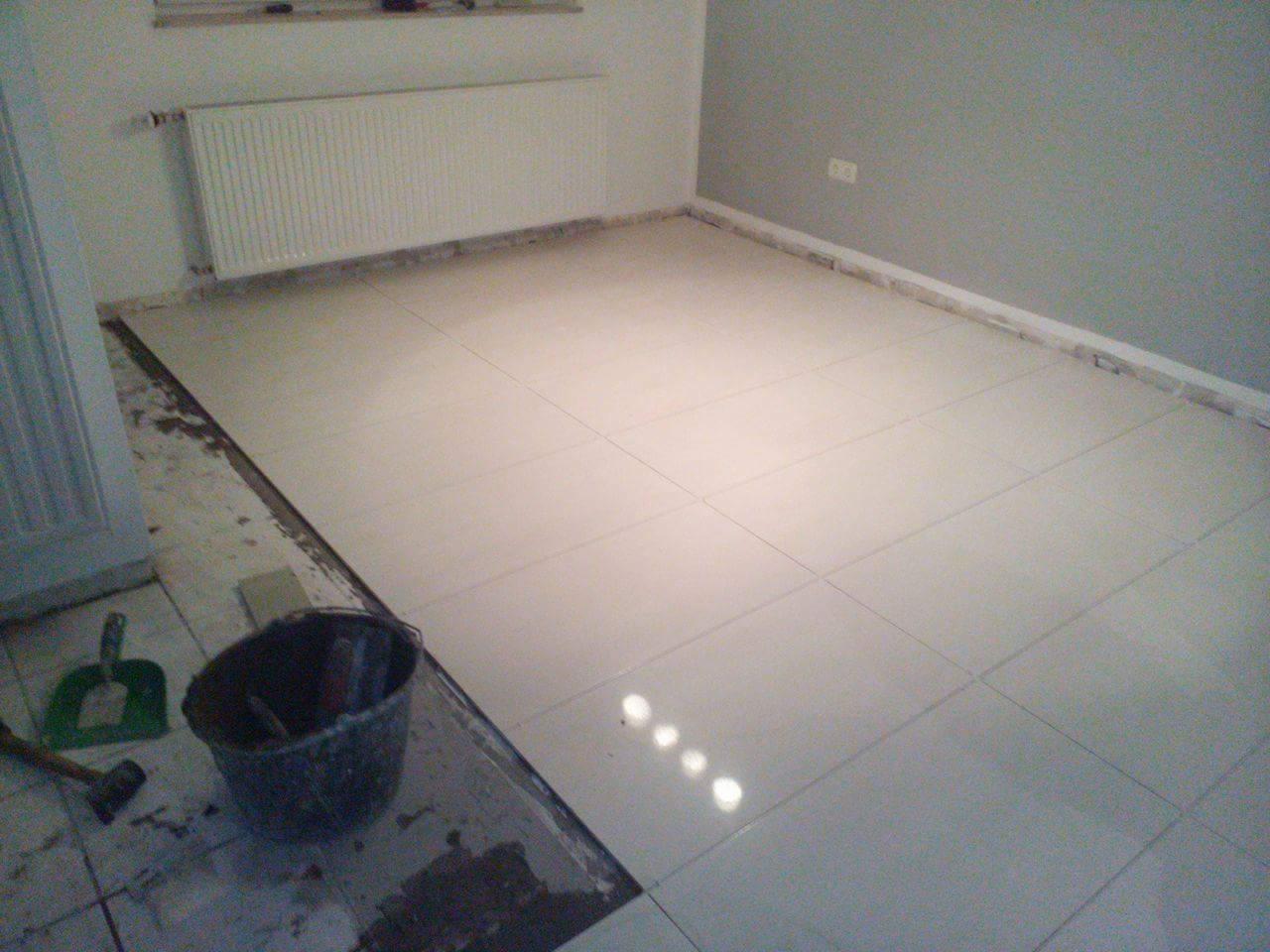 Ułożone jasne płytki podłogowe w pokoju, widoczny fragment podłogi przed położeniem płytek, wiadro z narzędziami i szpachelka w rogu.