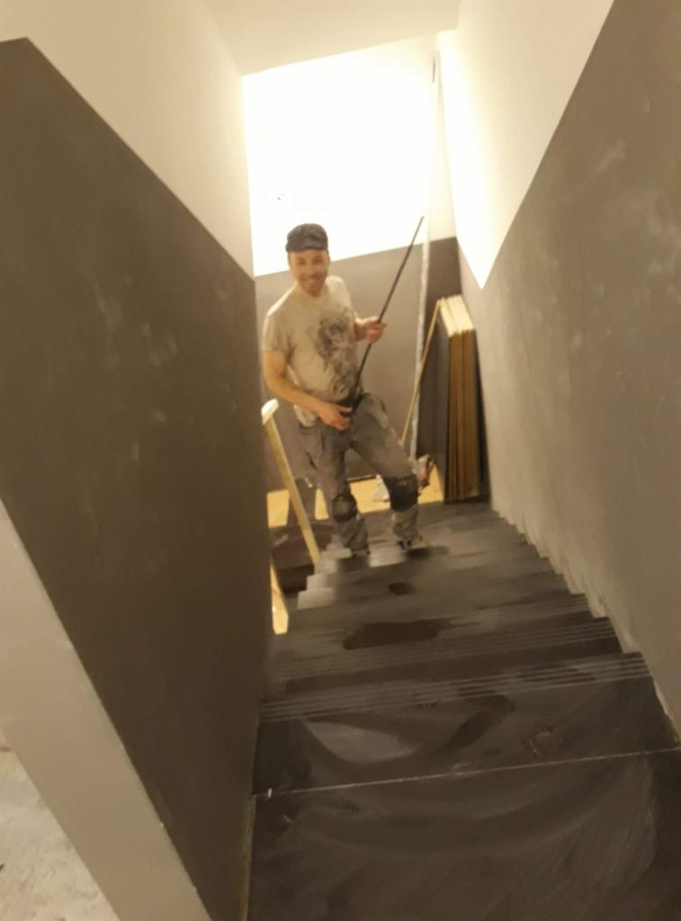 Pracownik w ubrudzonych farbą spodniach i czapce, stojący na schodach zabezpieczonych folią, z listwą w ręku, ściany pomalowane do połowy na szaro