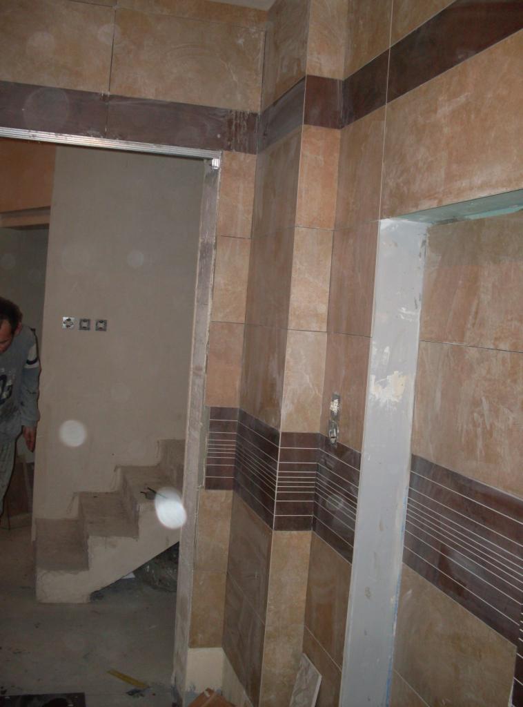 Wykończenie wnętrza z beżowymi i brązowymi płytkami, widoczne schody w surowym stanie, fragment osoby w szarej bluzie po lewej stronie kadru.