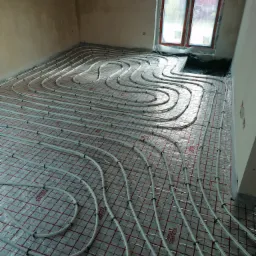 Układanie rur ogrzewania podłogowego na folii z siatką w surowym pomieszczeniu z oknem, widoczne złączki i napisy na folii.