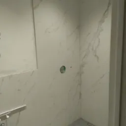 Wykończona łazienka z białymi płytkami imitującymi marmur, widoczne przyłącza hydrauliczne, odpływ liniowy w podłodze i otwór pod instalację elektryczną.