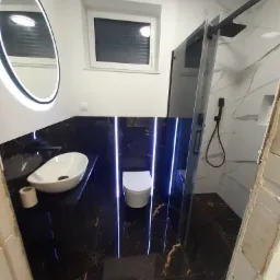 Nowoczesna łazienka z czarnym marmurem, owalną umywalką nablatową, podświetlanym lustrem LED i kabiną prysznicową typu walk-in z czarną armaturą.